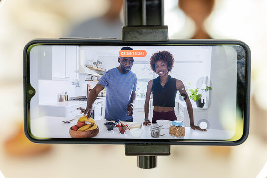 Diverse couple co-hosts preparing breakfast bowls at kitchen island with phone on tripod recording