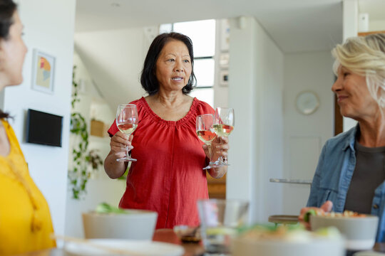 Diverse female friends listening while senior woman in red blouse holding two wine glasses at table