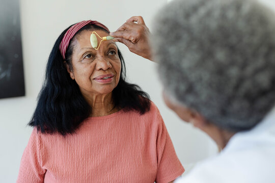 Senior African American women sitting at home, using jade roller on forehead, wearing coral top
