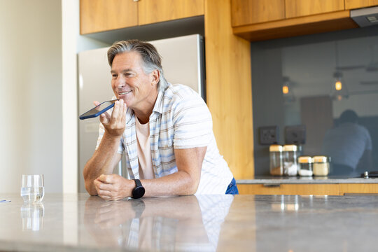 Mature man wearing striped shirt, leaning on stone counter speaking into smartphone in kitchen
