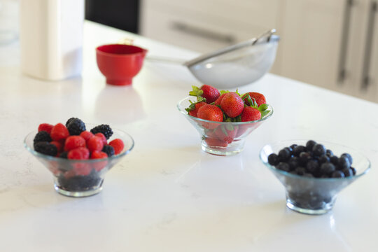 Clear glass bowl of strawberries is sitting on white marble counter with raspberries, blueberries