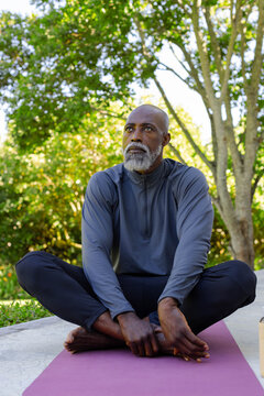 Senior African American man in sportswear sitting on yoga mat, yoga block on patio, holding feet