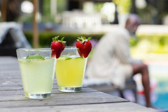 Two short clear glasses are sitting on slatted deck near pool, garnished with strawberries and mint