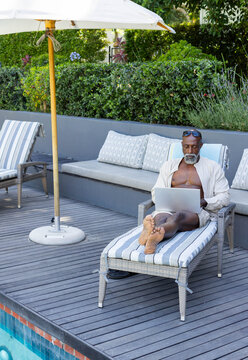Mature African man reclining on padded striped chaise lounge, using laptop at pool deck, copy space