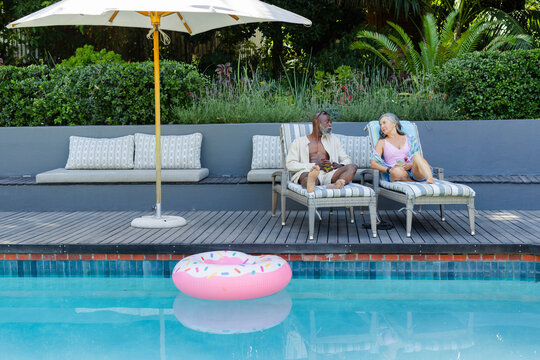 Couple sitting and conversing on striped lounge chairs on wooden deck by pool with pink float