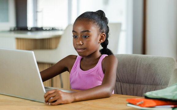 Child is studying on silver laptop at wooden table in kitchen with orange notebook and backpack