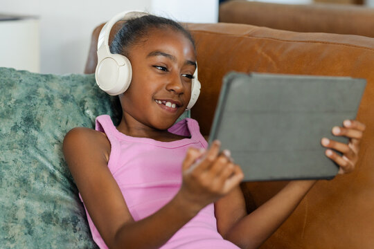 Gray tablet resting in landscape on brown leather sofa, with white headphones and green pillow