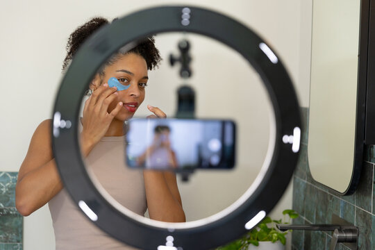 African American woman in beige tank applying blue under-eye patch at sink filming ring-light phone