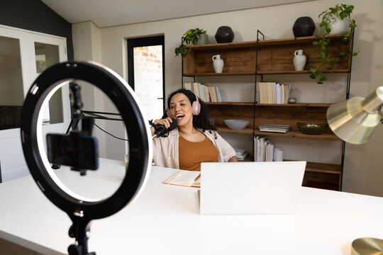 African American woman wearing headphones holding microphone and recording at studio with laptop