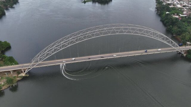 Cinematic drone shot of the Adomi Bridge arching over the Volta River in Akosombo, Ghana. Aerial view of West African infrastructure, river transport, and lush landscape.