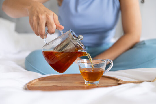 Glass teapot is pouring amber tea gently into clear glass cup on wooden tray on bed