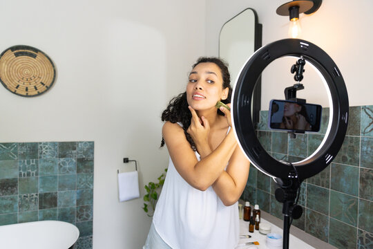 African American woman wearing white top applying gua sha recording phone with ring light at vanity