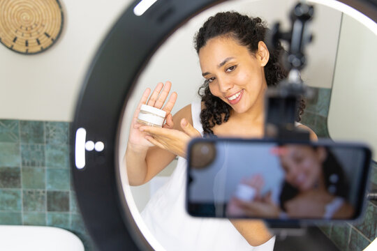 African American woman holding skincare jar in ring light, presenting to phone tripod at vanity
