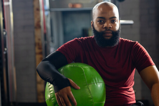 African American man sitting at gym holding bright green medicine ball while wearing maroon t-shirt