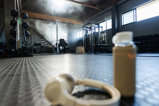 Over-ear headphones resting on warehouse gym floor, with water bottle, power rack, light haze