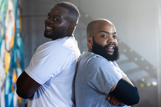 African American men standing back-to-back with arms crossed in gym warehouse with mural, staircase