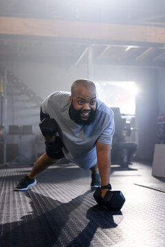 Man holding hex dumbbell rowing in plank on textured rubber floor wearing gray shirt shorts tracker