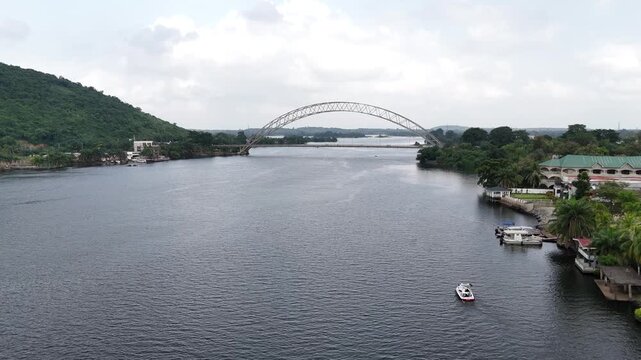Cinematic drone view of the Adomi Bridge crossing the Volta River in Akosombo, Ghana. Features the lush river banks and infrastructure of the West African landscape.