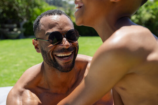 African American male friends laughing shirtless, leaning on garden lawn wearing round sunglasses