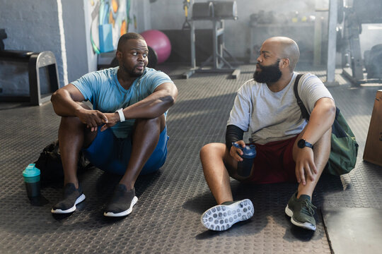 African American male friends resting and talking on gym floor wearing workout gear holding shakers
