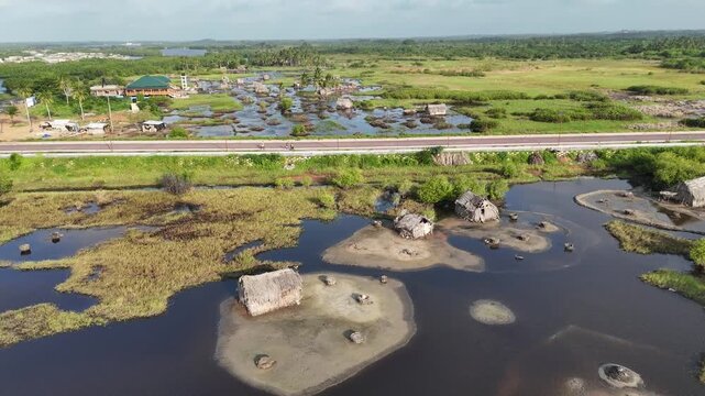 High-angle drone shot of a modern road splitting the lush marshlands of Ouidah, Benin. The footage shows traditional thatched-roof huts built over the water in a remote African landscape