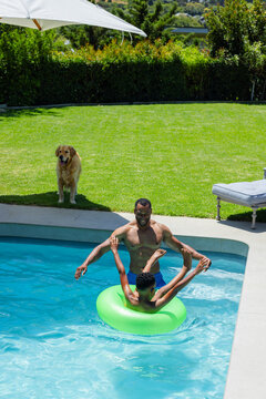 African American man and teenage boy holding hands, floating on neon green ring in backyard pool