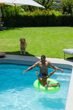 Father supporting child in sunny yard pool, shirtless dad in trunks holding green float and goggles