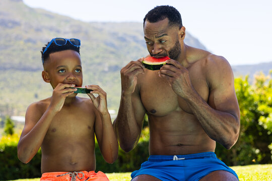 African American father and son eating watermelon wedges on grass at park wearing swim trunks