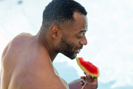 Shirtless mature man biting into watermelon slice by pool, holding rind with hand near sunlit face