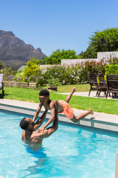 African American shirtless father holding son above water in backyard pool wearing orange trunks