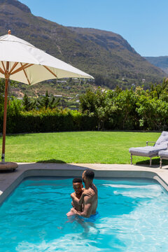 African American father holding son in backyard pool, wearing blue and patterned trunks, umbrella