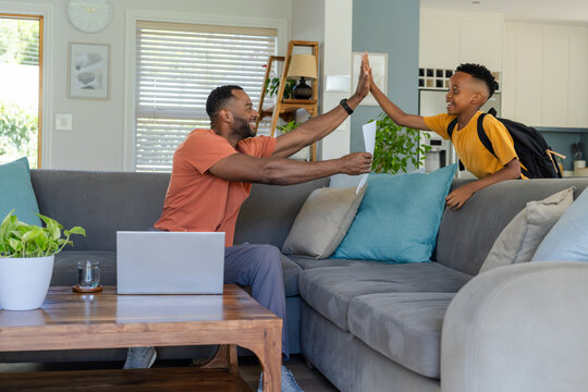 African American male father and child sharing high-five on sofa with backpack, paper and laptop