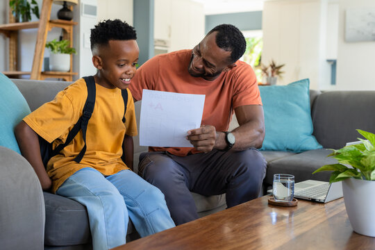 African American father and youth holding report showing letter A+ smiling with backpack on sofa