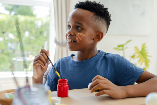 African American boy child painting at wood table by window wearing blue shirt holding yellow brush