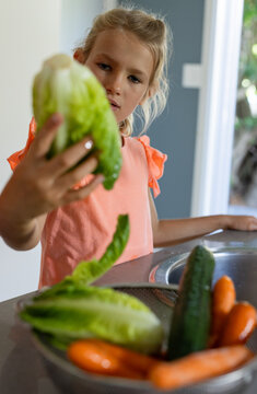 Female child holding romaine lettuce and rinsing at kitchen sink with colander wearing coral top