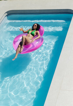African American woman reclining on pink float wearing green bikini, holding glass at private pool