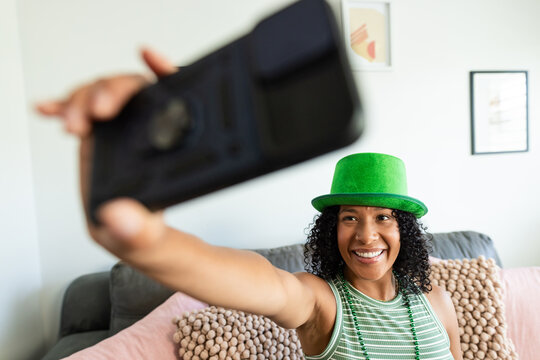 African woman sitting on couch taking selfie at home holding smartphone wearing green hat and beads
