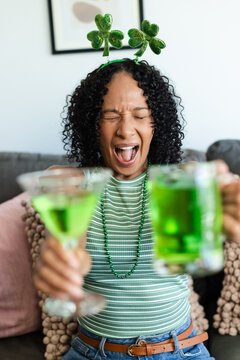 Adult African American woman with green headband holding martini glass, beer mug cheering on couch