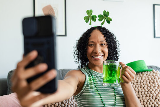 African American woman sitting on sofa at home wearing shamrock band holding phone and green drink