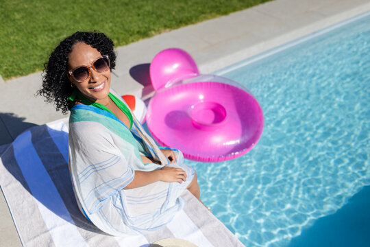 African woman sitting on towel at poolside wearing shades and cover-up, pink float, straw hat near