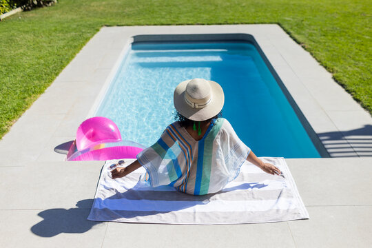 Woman sitting on white towel at backyard pool with pink inflatable ring wearing straw hat