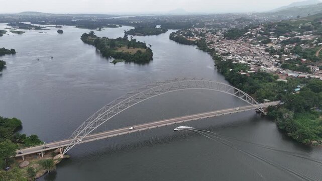 High-angle drone shot of the iconic Adomi Bridge in Atimpoku, Ghana. Cinematic view of the steel arch bridge crossing the wide Volta River with lush green banks and a small boat passing