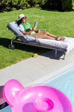African American woman reclining on lounge poolside in green bikini straw hat holding tablet
