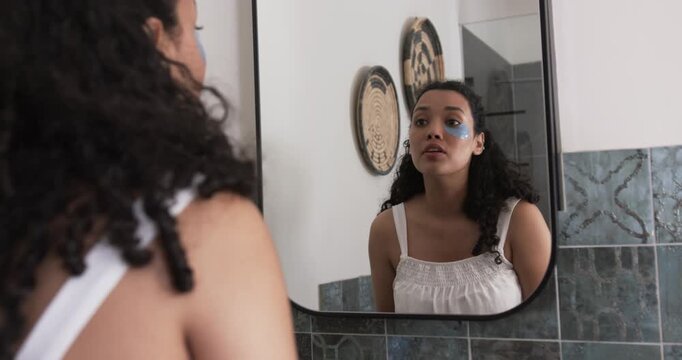 Woman checking skin, applying, peeling blue under-eye patches at bathroom mirror reducing puffiness