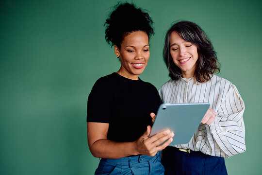 Two smiling women from diverse backgrounds collaborating and sharing ideas while using a portable digital tablet device against a green background
