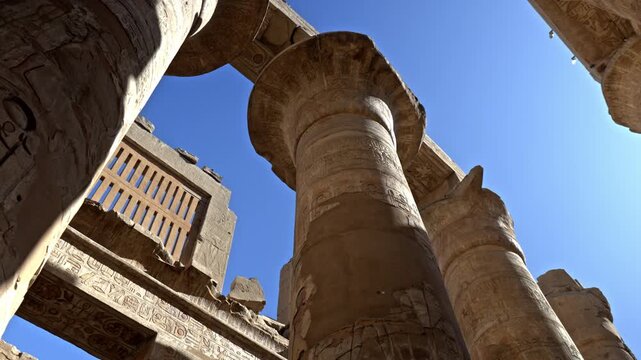 The composition highlights the horizontal stone architraves resting atop these pillars of Karnak in Luxor, framed against a deep blue sky. Sunlight illuminates the deep-relief hieroglyphic carvings.
