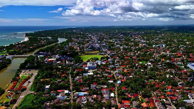 A drone captures an expansive aerial view of Matara's Nilwala River crocodile safari area, showing a sprawling town with houses, roads, lush greenery and glimpses of the coastline under a blue sky.