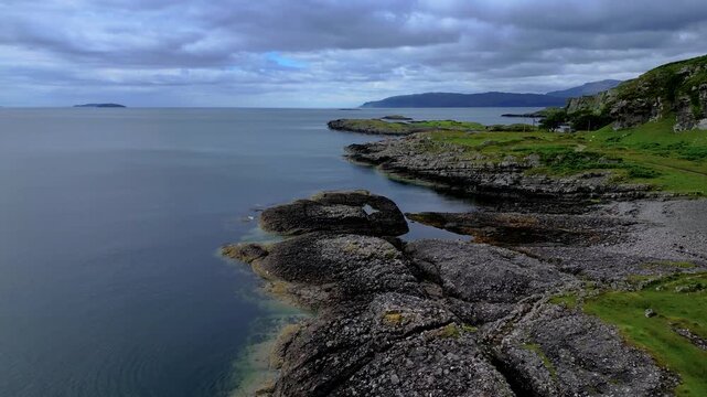 Aerial drone flight over rugged rocky coastline of Isle of Mull with green hills and calm sea. Dramatic Scottish island landscape under cloudy sky. Remote and wild Hebrides coastal scenery Scotland.