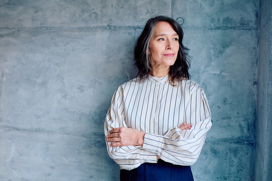 Mature woman with gray hair crossing arms, looking away thoughtfully against a concrete wall, embodying professional confidence and contemplation