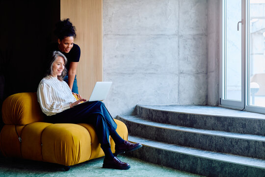 Two diverse business women collaborating, using a laptop and discussing work in a contemporary office setting, sharing ideas
